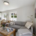 Wohnbereich mit Sofa, Esstisch und Küchenzeile. Fenster mit Blick auf Außenbereich.