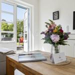Ein Wohnzimmer mit Holztisch, Blumen und TV. Durch die Tür ist ein Garten sichtbar.