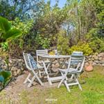 Weiße Gartenmöbel um einen Tisch mit Wein und Obst. Steinmauer und grüne Pflanzen im Hintergrund.