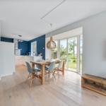Ein Essbereich mit Holztisch und Stühlen, Küchenbereich und TV-Wand mit Flur im Hintergrund.