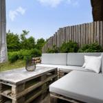Terrasse mit grauem Sofa und Tisch aus Paletten. Hintergrund: Holzzaun und grüne Pflanzen.