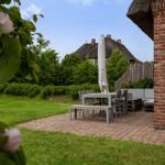 Terrasse mit Tisch, Stühlen und Sitzbank neben Backsteinmauer. Grasfläche und Rosenbusch im Vordergrund.