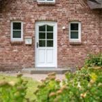 Haus mit rotem Ziegelsteinfassade, weißer Tür und Fenstern, gepflegter Garten mit Blumen.
