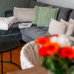 Ein grauer Sofa mit Kissen und einer Decke. Ein Tisch und Blumen sind im Vordergrund.