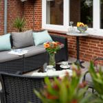 Terrasse mit Sitzgruppe, Blumen und Obstschale. Holzdielen und Backsteinmauer.