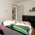 Schlafzimmer mit Bett, Schrank und Wandregal. Tür führt in Wohnbereich.