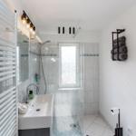 Badezimmer mit Dusche, Waschbecken und Toilette. Fenster, Handtuchhalter und Heizung sichtbar.