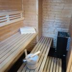 Sauna mit Holzbank, Steinkachelofen und Eimer mit Löffel.