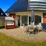 Terrasse mit Strandkorb, Tisch, Stühlen und Liege. Haus mit Glasfront und Balkon.