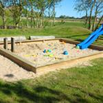 Holzrahmen-Sandkasten mit blauer Rutsche und Spielzeug im Garten.