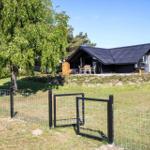 A house with a dark roof and terrace stands on a green lawn behind a fence.