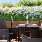 Terrace with table and chairs, view of green hedge with white flowers.