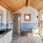 Badezimmer mit Dusche, Waschbecken und Toilette. Holzdecke und Fenster mit Holzrahmen.