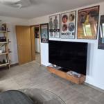 Wohnzimmer mit Fernseher, Filmplakaten und Holzregal. Fliesenboden und Tür im Hintergrund.