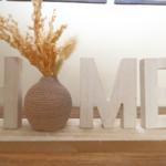 Wooden letters spelling HOME and a vase with dried plants on a wooden surface.