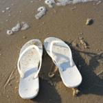 Weiße Flip-Flops liegen am Strand mit Sand und Schaum.