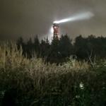 Roter Leuchtturm mit Lichtstrahl durch Nacht, umgeben von Bäumen und Sträuchern.