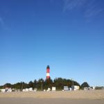 Roter Leuchtturm mit weißer Markierung, umgeben von Strandkörben und Bäumen unter blauem Himmel.