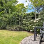 Garten mit Rasen, Steinterrasse und Gartenmöbeln. Hintergrund: dichte Hecke und Bäume.