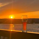Ein Bierglas steht auf einer Terrasse vor einem See bei Sonnenuntergang.