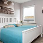 Schlafzimmer mit weißem Bett, blauer Bettdecke und Fenster mit Blick auf Strand.