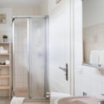 Badezimmer mit Dusche, Waschbecken und Holzregal mit Handtüchern und Pflanzen.