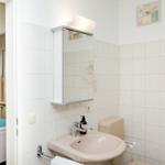 Badezimmer mit Waschbecken, Spiegel und Toilette. Blick in das Schlafzimmer mit Bett.