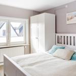 Schlafzimmer mit weißem Bett, Kleiderschrank und Fensterblick auf Dächer.