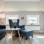 Living room with sofa, armchair, table, and TV. Wooden floor and two windows.