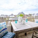 Terrasse mit Holztisch, Stühlen und Fischmotiv-Kissen. Blick auf Wohnhäuser und Himmel.