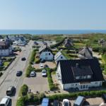 Haus mit dunklem Dach, umgeben von Grünflächen und Parkplätzen. Im Hintergrund liegt das Meer.