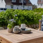Holztisch mit Tassen, Tellern und Blumen im Garten. Hintergrund: grüne Hecke und Häuser.