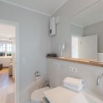 Badezimmer mit Waschbecken, Spiegel und Toilette. Durchgang zu Schlafzimmer mit Holzboden.