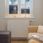 Ein Wohnbereich mit Sofa, Fußhocker und zwei Lampen am Fenster.