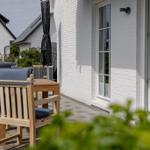 Terrasse mit Holztisch und Stühlen vor weißer Hauswand mit Fenstern.
