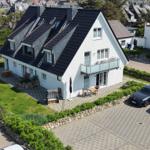 Weißes Haus mit dunklem Dach, Terrasse und Garten. Parkplatz mit zwei Autos.