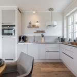 Moderne Küche mit weißer Einbauküche, Edelstahlspüle und Holzboden. Fenster und Deckenleuchten sorgen für Licht.