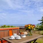 Terrasse mit Tisch, Stühlen, Blumen und Gebäck. Blick auf Meer und Himmel.