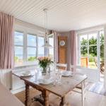 Esszimmer mit Holztisch, Fenstern zum Meer und Terrasse. Holzdecke, rosa Vorhänge.