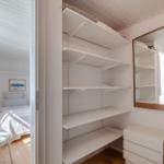 Schrank mit weißen Regalen und Spiegel. Blick in Schlafzimmer mit Bett und Nachttisch.
