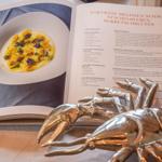 Offenes Kochbuch mit Rezept und einem goldenen Käsehobel vor einem Teller.