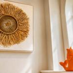 Wandbild mit Strohdekor neben einem Fenster mit orangefarbener Fischfigur.