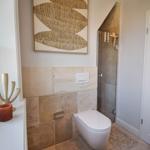 Badezimmer mit Toilette, Dusche und Fenster. Wandverkleidung aus Stein und Kunstwerk an der Wand.