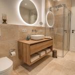 Badezimmer mit Dusche, Waschtisch und Toilette. Holzunterschrank und runder Spiegel.