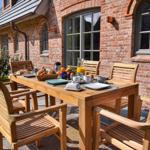 Holztisch mit Stühlen auf Terrasse vor Backsteinhaus mit Frühstückszubehör.