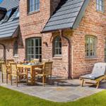Terrasse mit Holztisch, Stühlen und Liegestuhl vor Backsteinhaus mit Dachgauben.