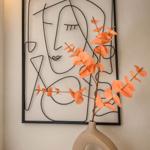 Wanddekoration mit abstraktem Gesicht und orangefarbener Blumenvase auf Marmorplatte.