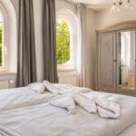 Schlafzimmer mit Bett, Kleiderschrank und zwei Bogenfenstern mit Blick auf Grün.