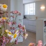 Küche mit weißen Schränken und Fenster mit Jalousie. Vordergrund: Blumenstrauß mit Kamillen und rosa Blüten.