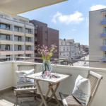 Balkon mit Tisch, zwei Stühlen und Blumen. Blick auf Straße und Gebäude.